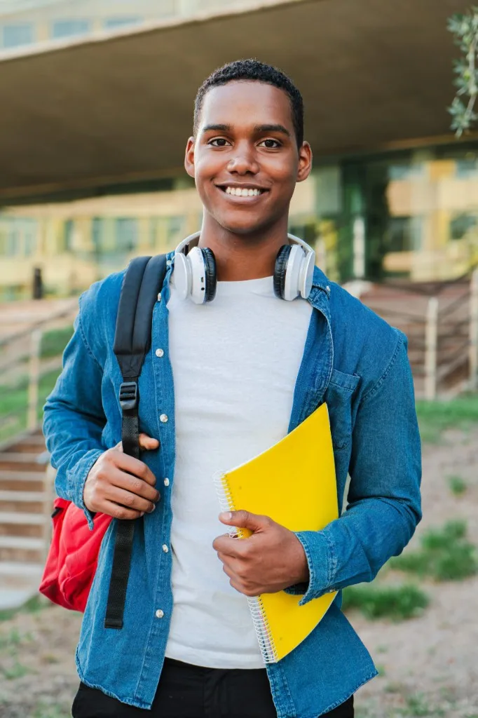 A smiling student with a backpack and notebook standing outside a campus building